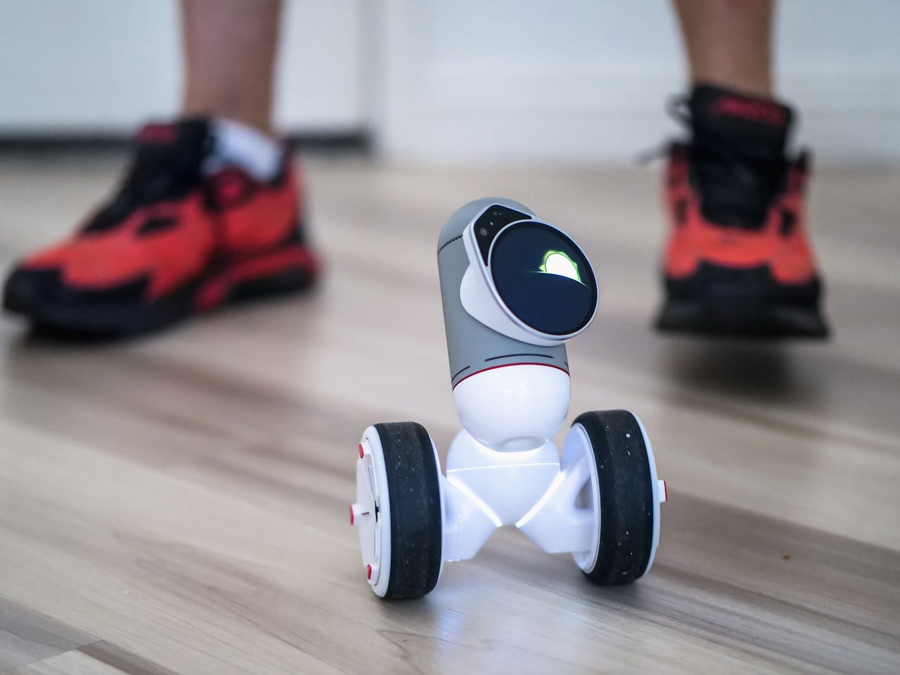 Close-up of a two-wheeled mini robot on a wooden floor with sneakers in background.